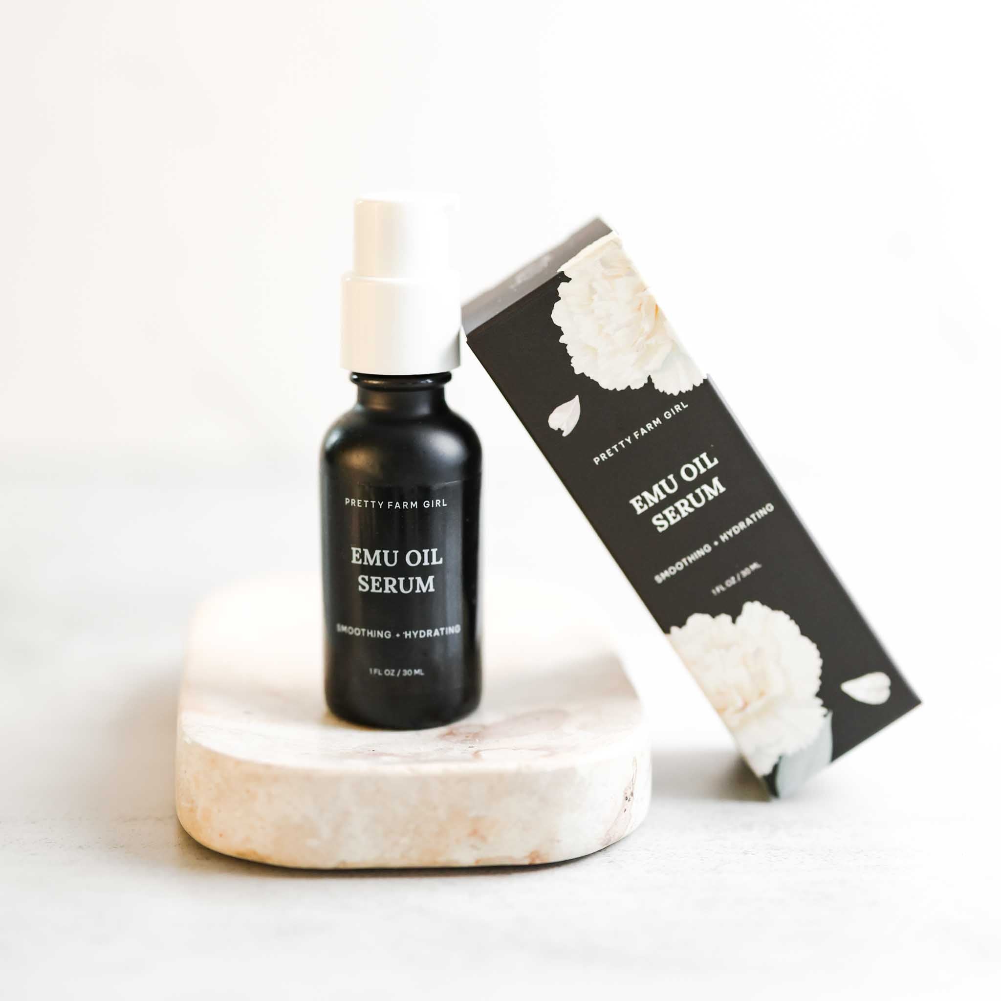 Emu Oil Serum in a black bottle with a white pump, displayed on a stone surface alongside its floral box packaging.