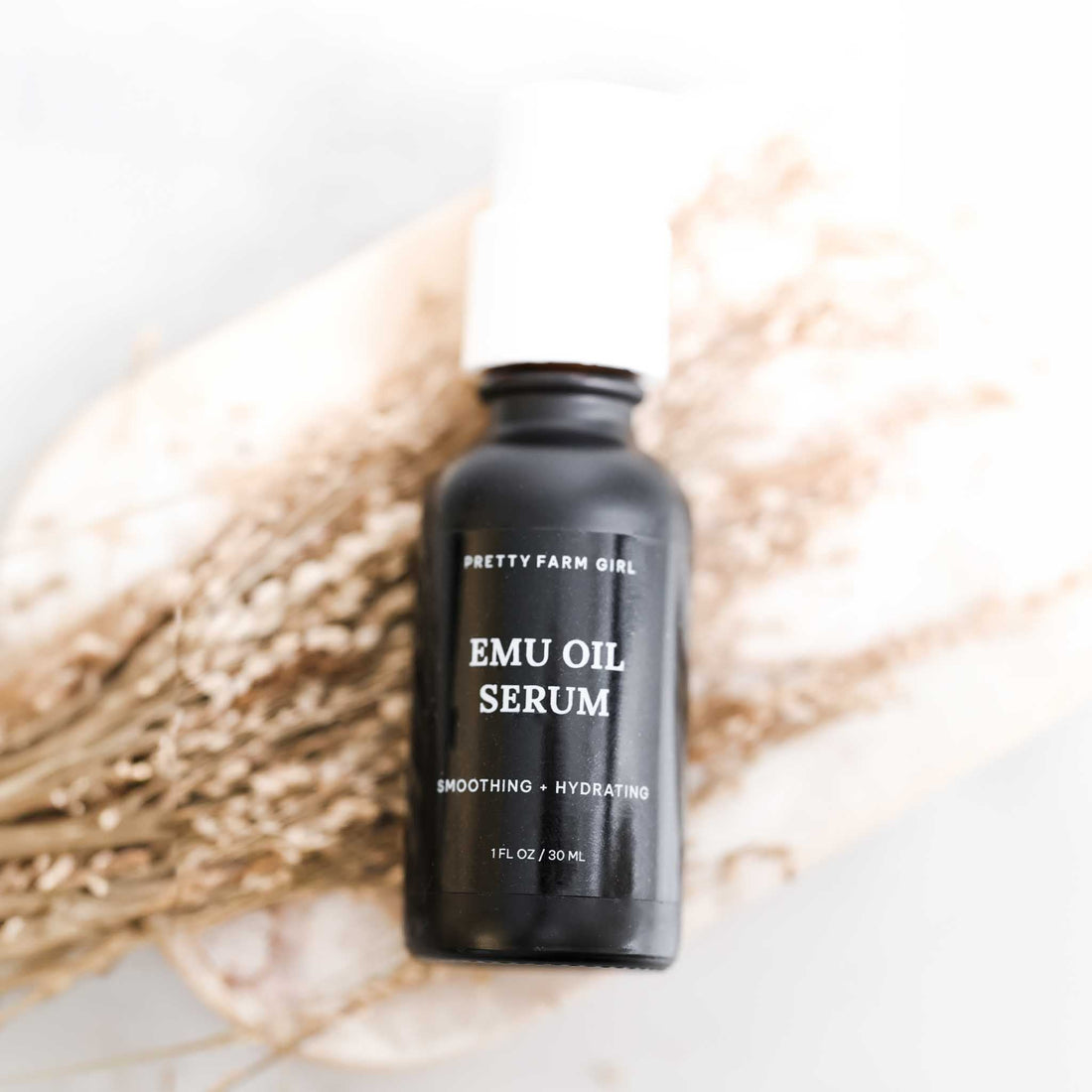 Emu Oil Serum in a sleek black bottle, 1 fl oz, labeled "Smoothing - Hydrating," set against a soft, natural background.