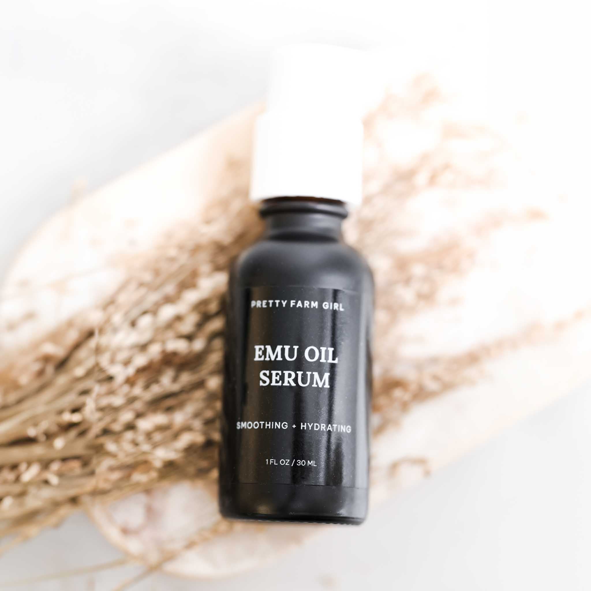 Emu Oil Serum in a sleek black bottle, 1 fl oz, labeled "Smoothing - Hydrating," set against a soft, natural background.