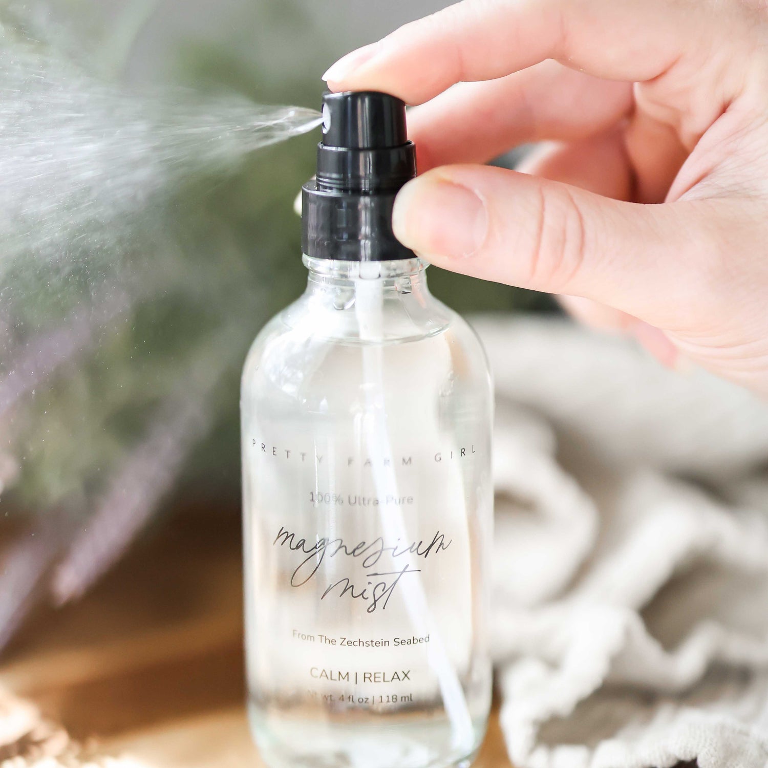 Magnesium Mist Body Spray in a clear bottle with a spray nozzle, being sprayed from a hand, on a wooden surface.