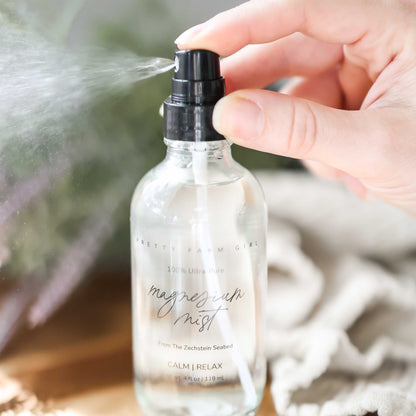 Magnesium Mist Body Spray in a clear bottle with a spray nozzle, being sprayed from a hand, on a wooden surface.